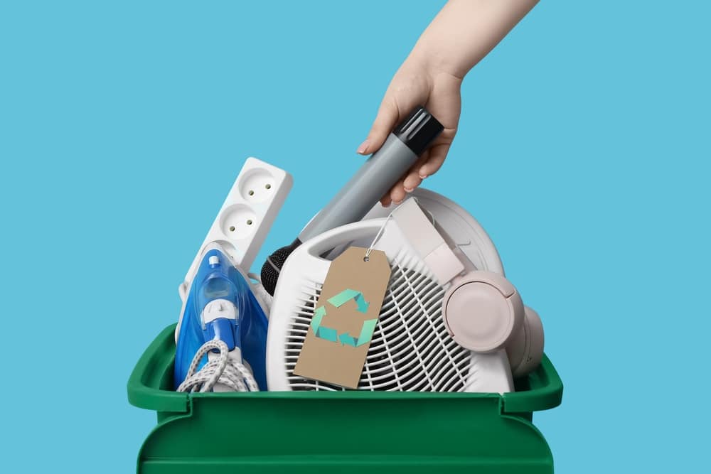 descarte eletrodomesticos reciclagem Imagem de uma pessoa colocando utensílios domésticos em uma caixa de reciclagem verde, incluindo um ferro, uma extensão elétrica, um fone de ouvido e um microfone, promovendo descarte responsável de lixo.
