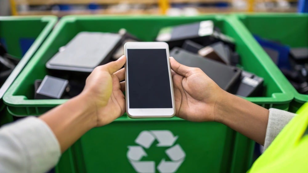 Pessoa segurando smartphone ao lado de uma lixeira de reciclagem cheia de eletrônicos usados, destacando a importância da reciclagem de eletrônicos.