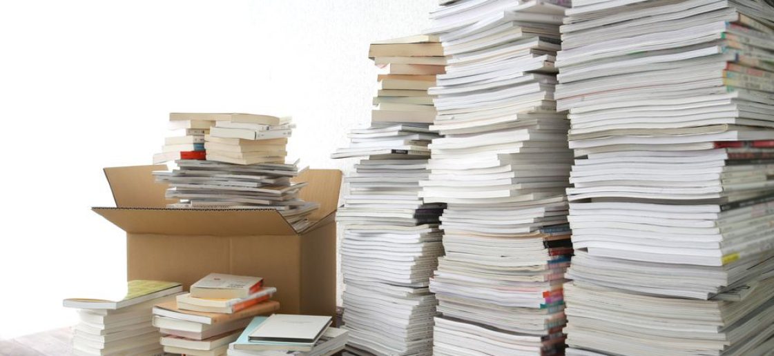 Pilhas de materiais antigos ao lado de uma caixa aberta, sugerindo a dúvida sobre onde descartar livros escolares acumulados.