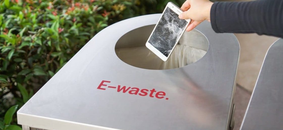 Pessoa descartando um celular quebrado em lixeira identificada como e-waste, voltada à reciclagem de eletrônicos.